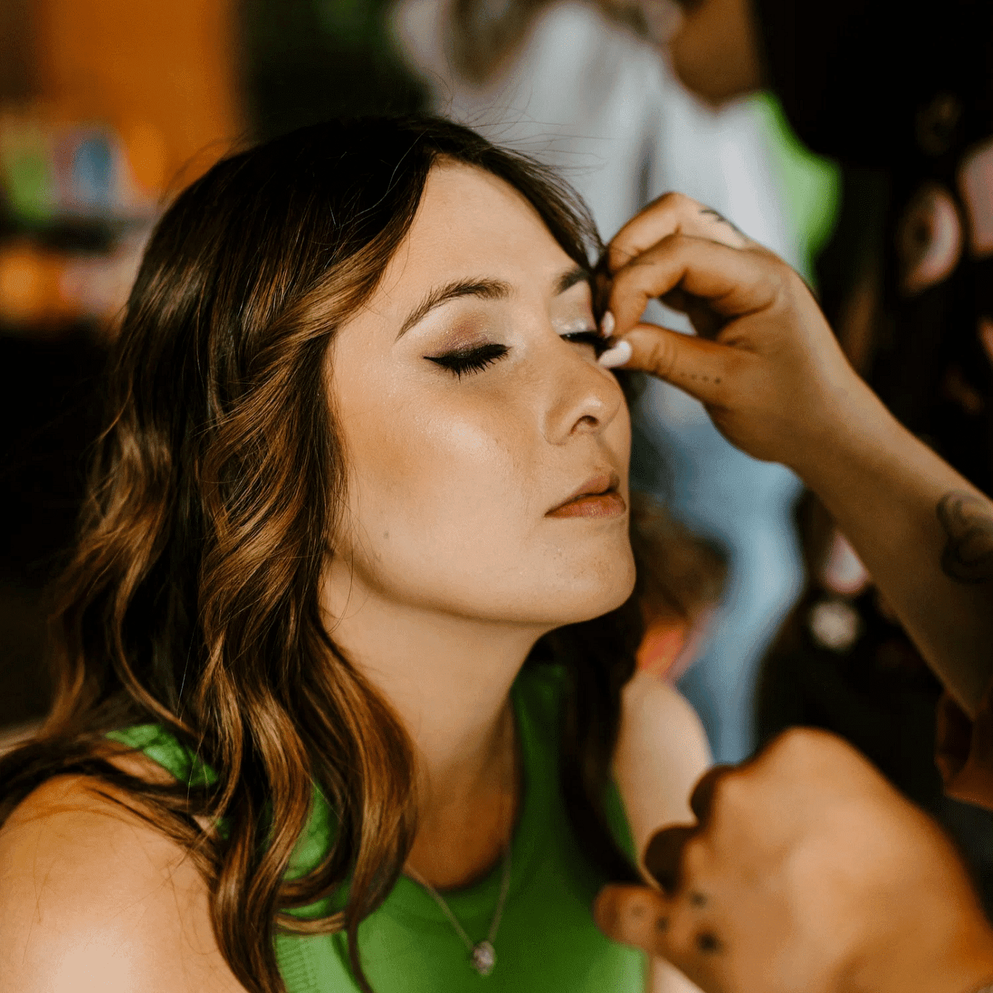 Woman in a green top getting makeup applied, eyes closed in a relaxed setting.