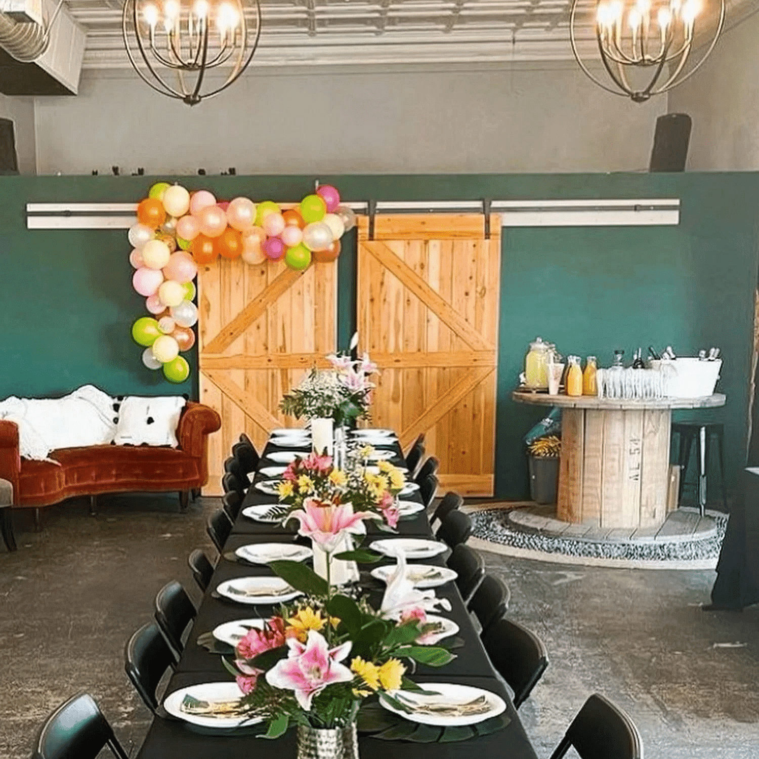 Long table set for a party with colorful flowers and balloons, rustic decor, and hanging chandeliers.