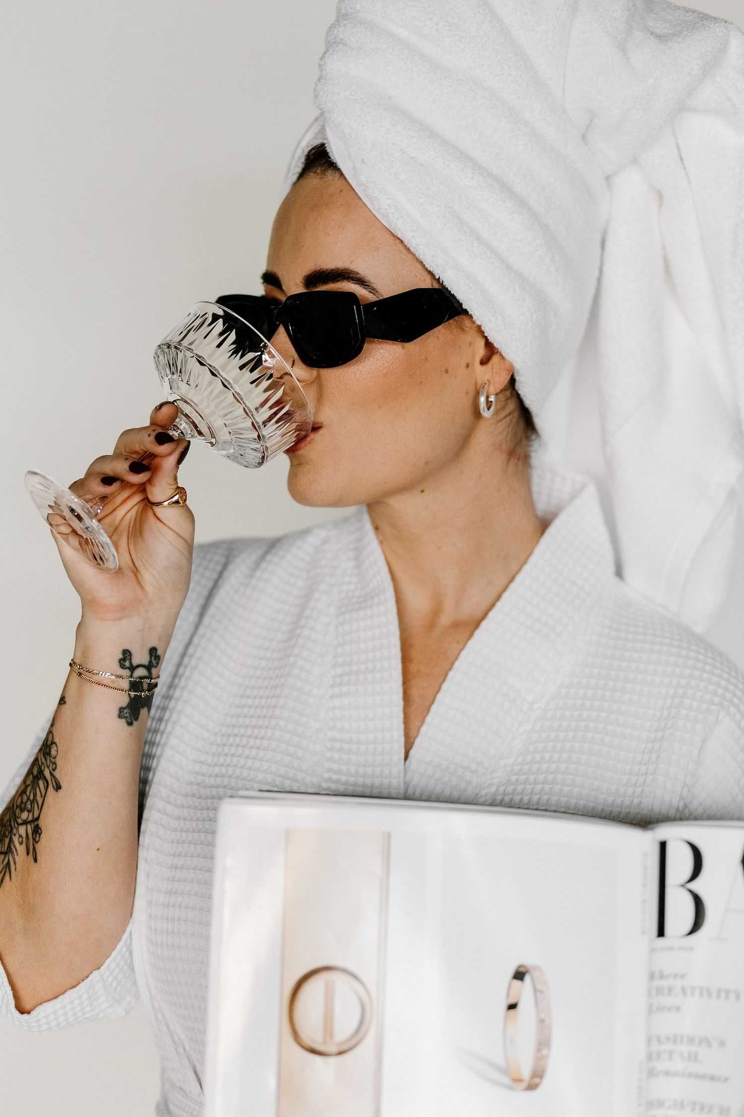 Woman in bathrobe and towel sipping from glass while holding a magazine, wearing sunglasses.