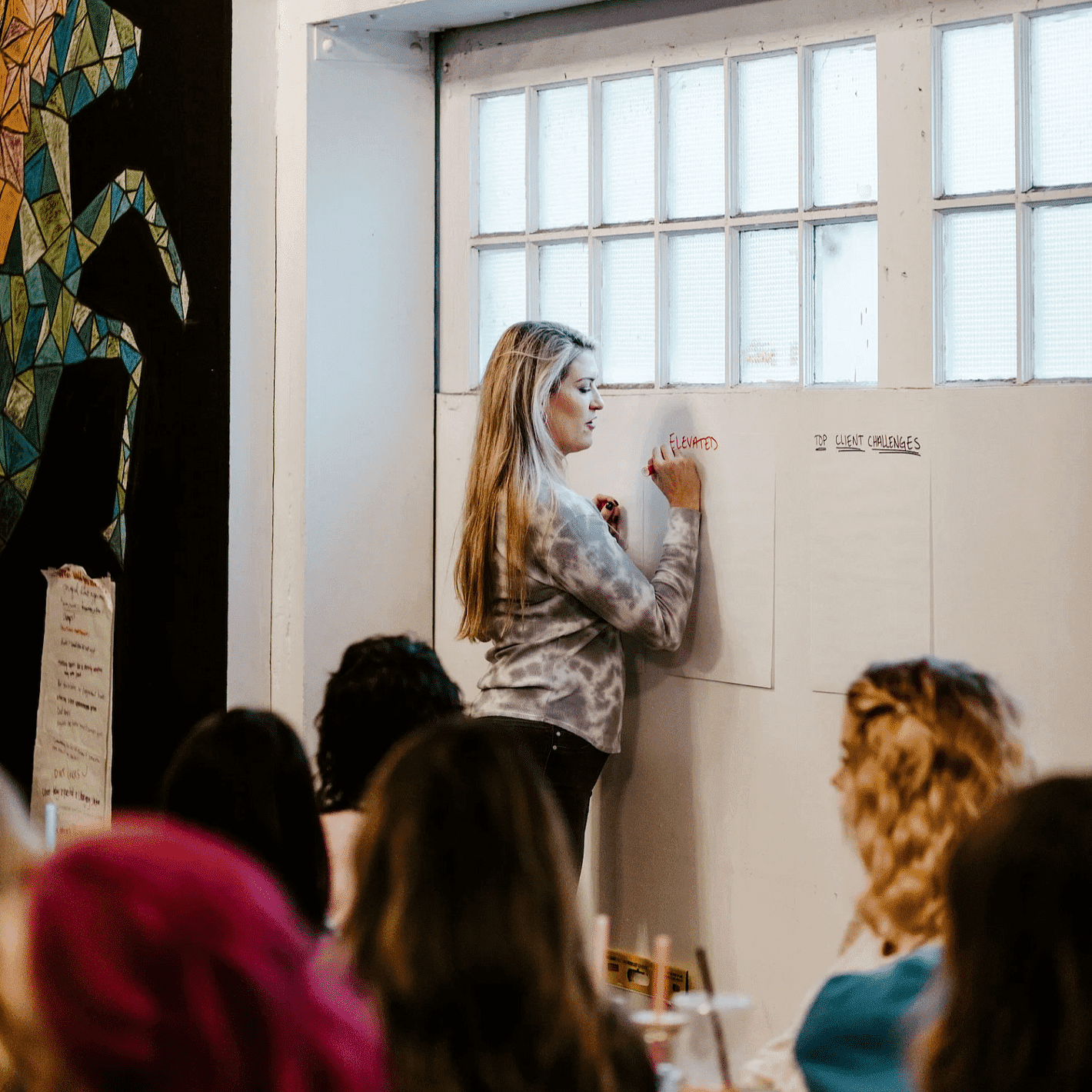 Woman presenting to a seated audience in a creative workspace with colorful mural and Exit sign.